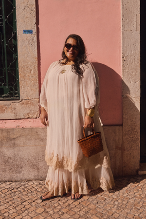 A woman in a flowing white dress, sandals, and sunglasses stands against a pink wall, smiling in the sunlight while holding the Hayden - Shell woven handbag.