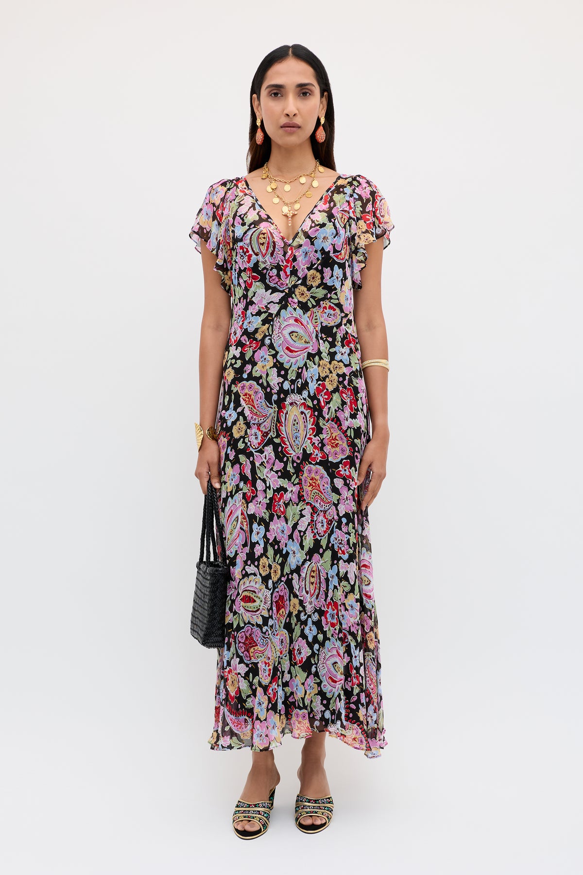 A woman with straight dark hair stands against a white background, wearing the Bexley - Cara Floral Black dress with short sleeves, accessorized with gold jewelry, black handbag, and sandals.