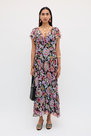 A woman with straight dark hair stands against a white background, wearing the Bexley - Cara Floral Black dress with short sleeves, accessorized with gold jewelry, black handbag, and sandals.