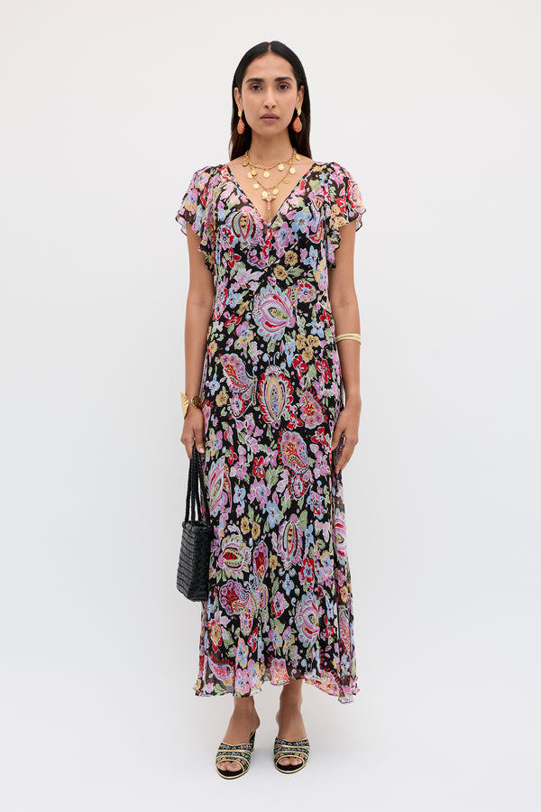 A woman with straight dark hair stands against a white background, wearing the Bexley - Cara Floral Black dress with short sleeves, accessorized with gold jewelry, black handbag, and sandals.
