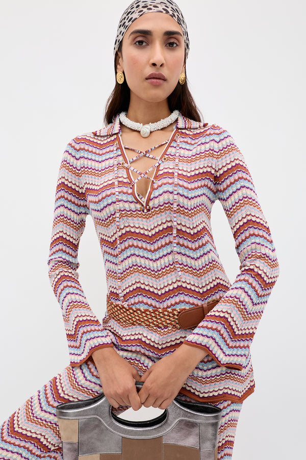 Wearing the Brody - Maria Chevron Multi, a vibrant zigzag-patterned outfit, this woman styles her vintage-inspired look with a lace-up V-neck top, accessorized with a headscarf, shell necklace, and gold earrings. She confidently poses against a plain background while holding a metallic handbag.