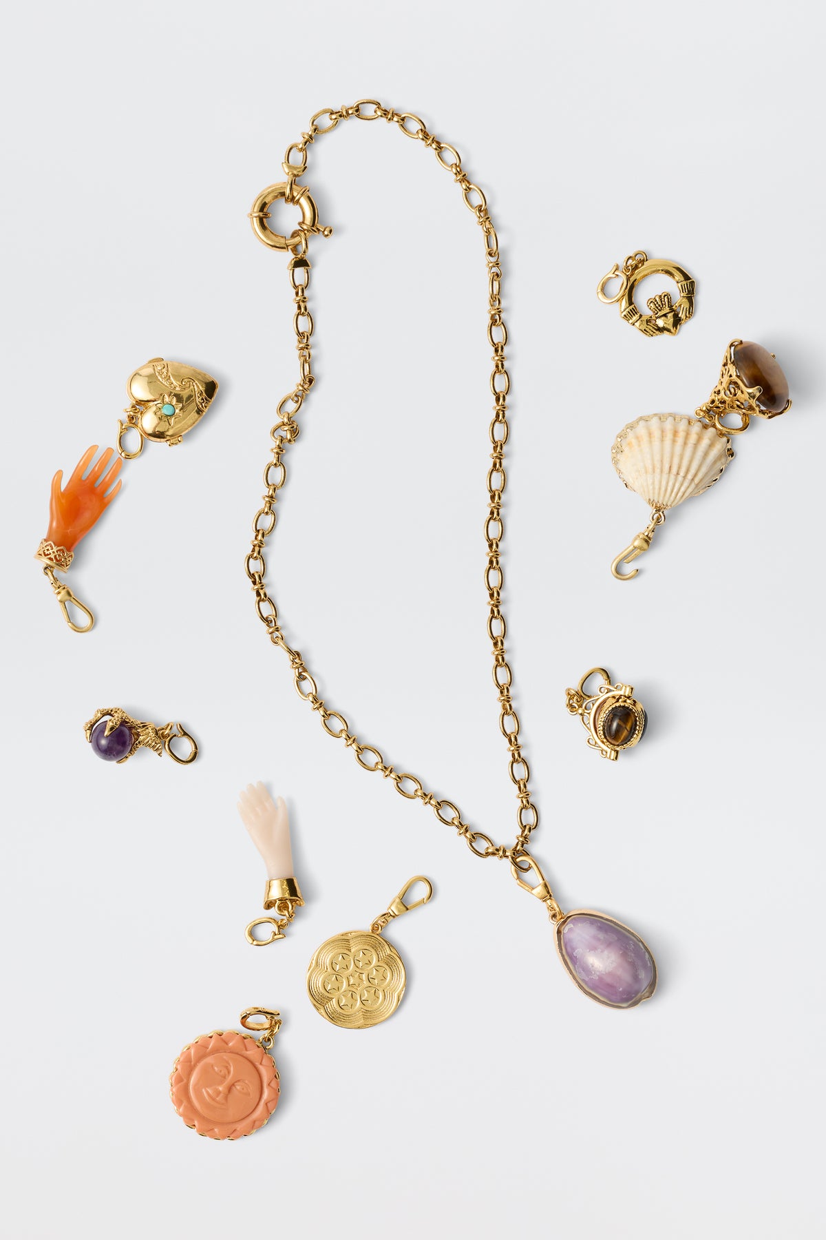 The Maella - Gold necklace features a gold chain with a purple gemstone pendant, surrounded by 18k gold-plated charms—hands, shell, hat, and engraved medallions—beautifully displayed on a white background.