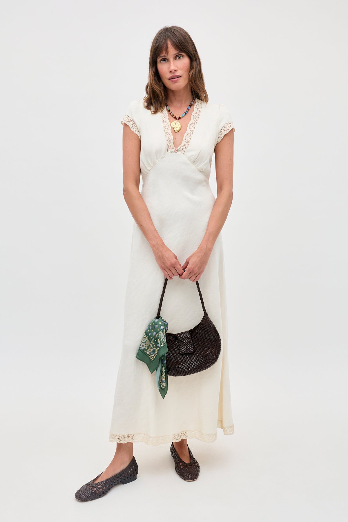 A woman in the Clarice - Cream vintage nightdress-style dress with lace trim stands holding a dark woven handbag with a green scarf, wearing flat brown shoes and layered necklaces against a plain white background.