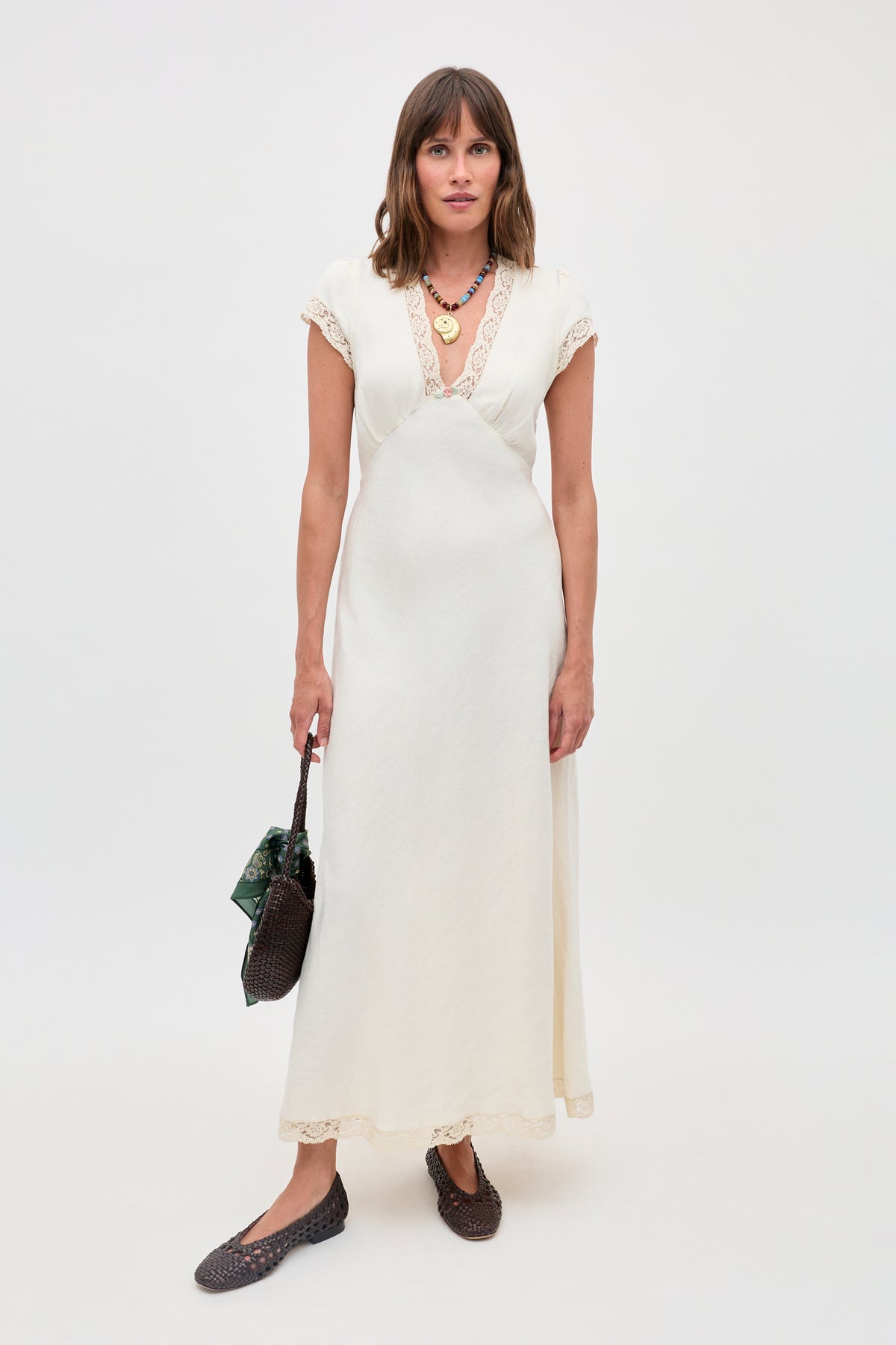 A woman stands against a plain white background wearing the Clarice - Cream vintage nightdress with lace trim, dark woven flats, layered necklaces, and holding a dark woven handbag adorned with a green scarf.