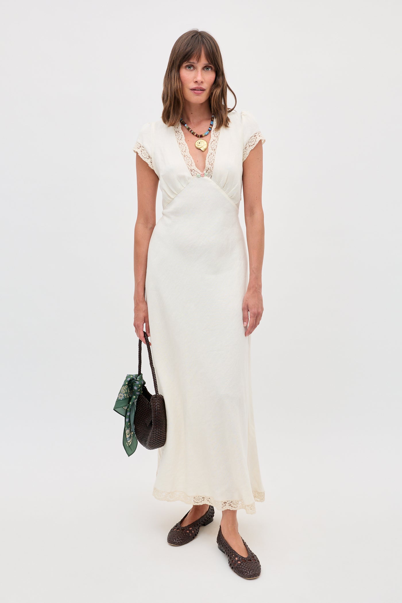 A woman poses against a white background in the Clarice - Cream vintage nightdress with lace trim, accessorized with a dark woven handbag featuring a green scarf, layered necklaces, and dark shoes.