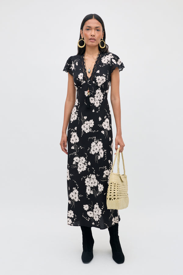 A woman wearing the Clarice - Daisy Field Black dress, paired with black boots, gold hoop earrings, a long necklace, and holding a light-colored woven handbag, stands against a plain white background.