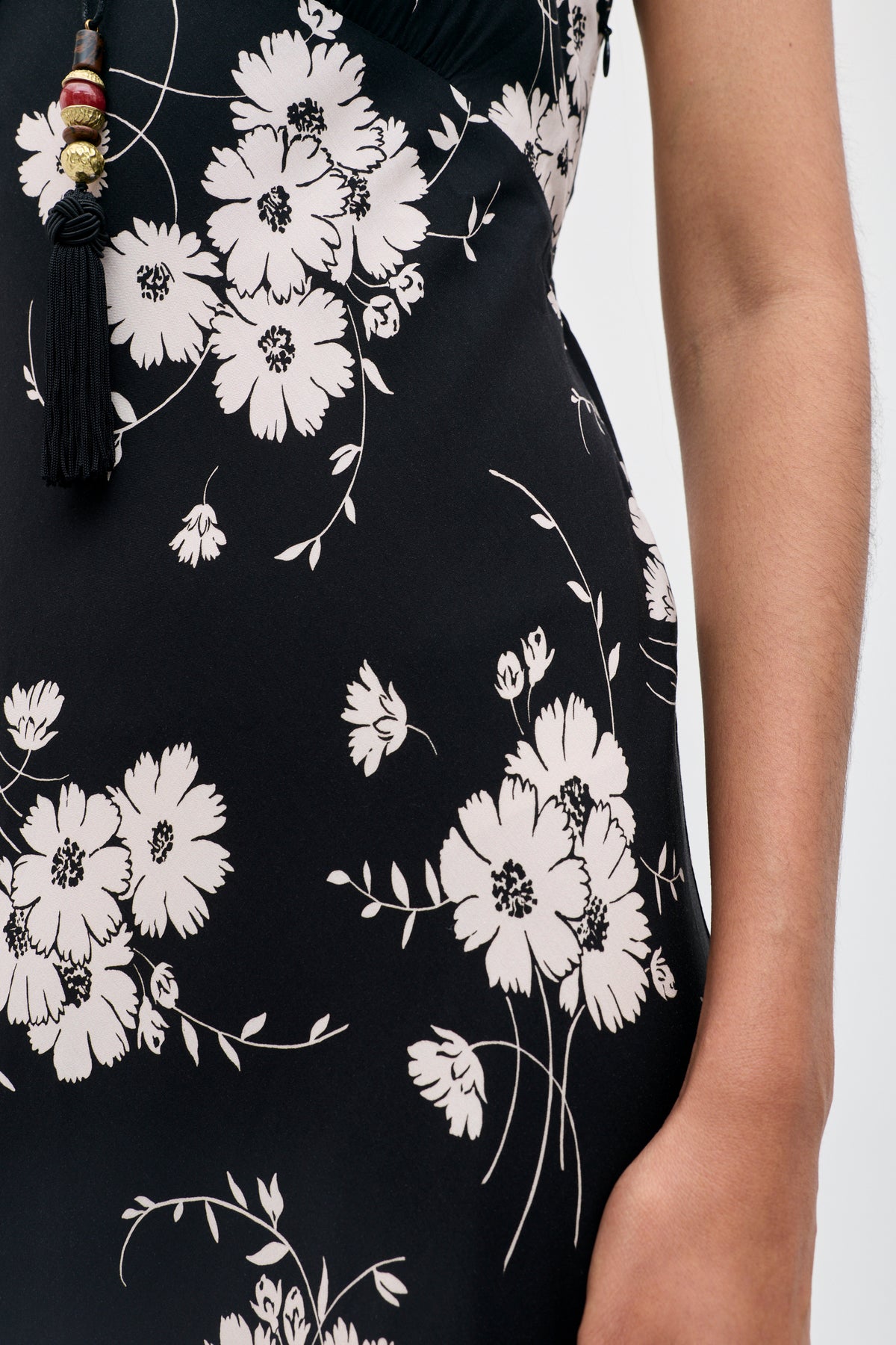 A close-up of a person wearing the Clarice - Daisy Field Black dress, featuring white floral patterns and a tassel necklace. The person’s arm is visible on the right side of the image.