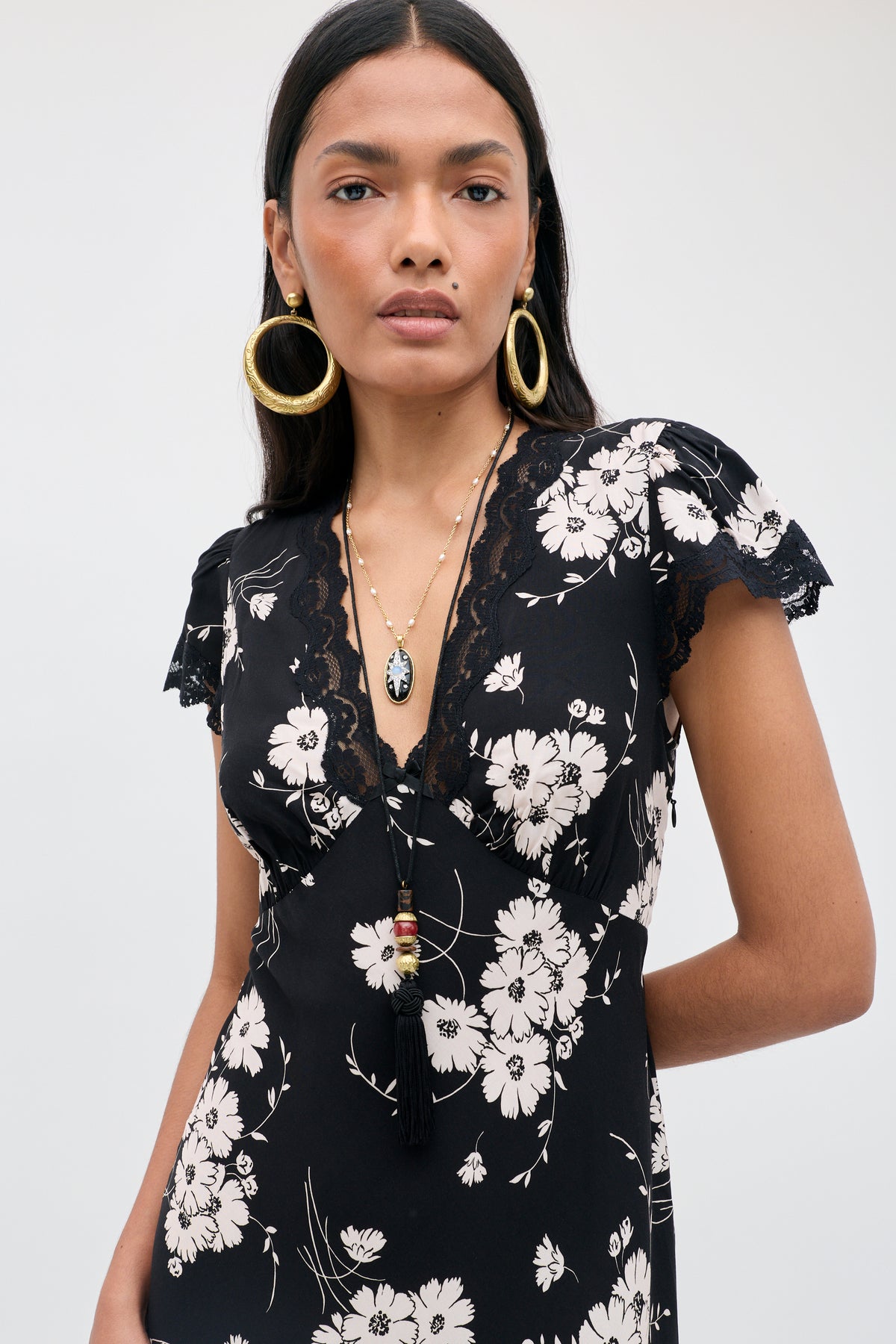 A woman wearing the Clarice - Daisy Field Black dress, which features a black base with a white daisy floral print and delicate lace trim on the sleeves and neckline. She accessorizes with large gold hoop earrings and layered necklaces, standing against a plain light background.
