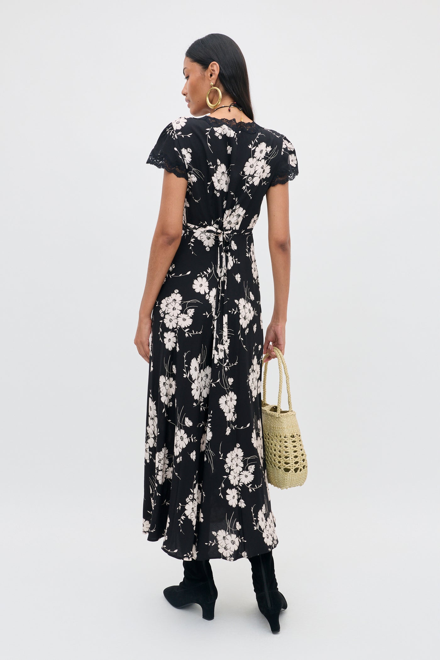 A woman stands facing away, wearing the Clarice - Daisy Field Black dress, paired with black boots, large hoop earrings, and holding a woven beige bag. Her hair is straight and dark, set against a plain white background.
