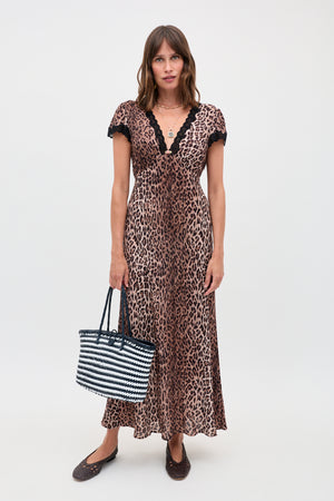 A woman with straight brown hair and a neutral expression stands against a plain background, wearing the clarice dress leopard brown with black lace trim, holding a striped woven handbag, and wearing dark flat shoes.