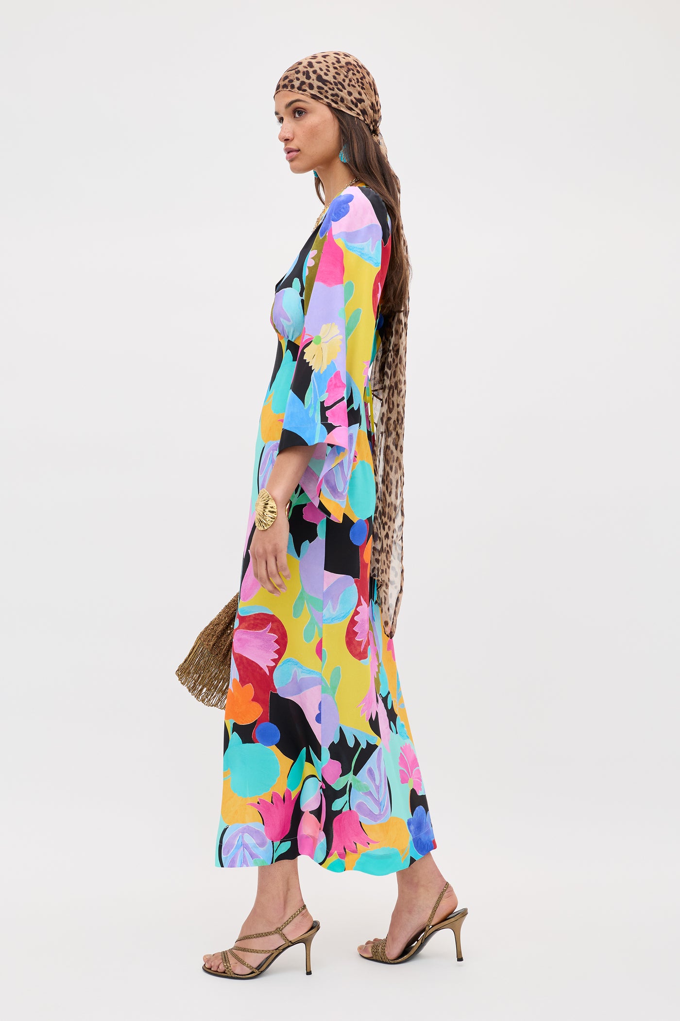 Woman in a dramatic Dahlia - Abstract Tulip Black silk crepe de chine maxi dress with bold sleeves, styled with a brown leopard-print headscarf, gold sandals, and a woven bag, standing against a plain white background.