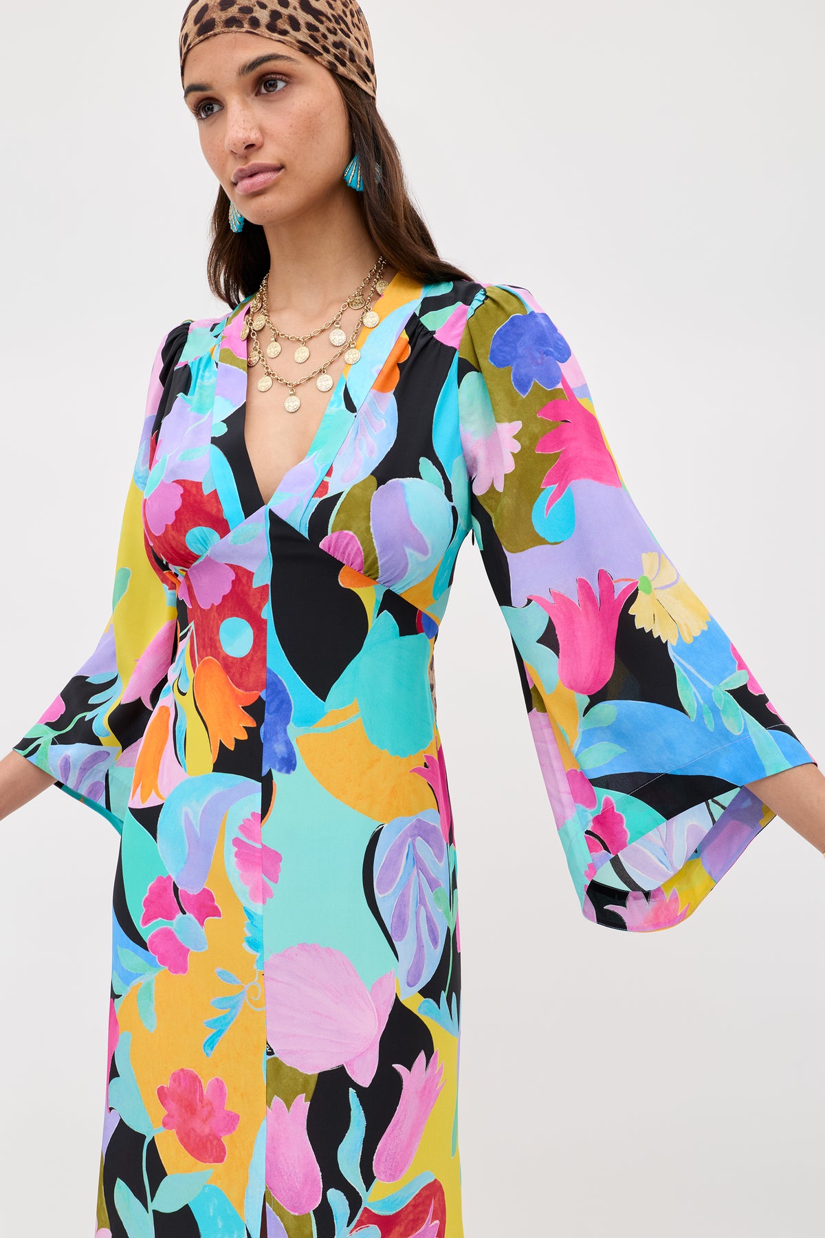 A woman wears the Dahlia - Abstract Tulip Black dress with dramatic sleeves, layered necklaces, turquoise earrings, and a leopard-print headscarf. The background is plain white.