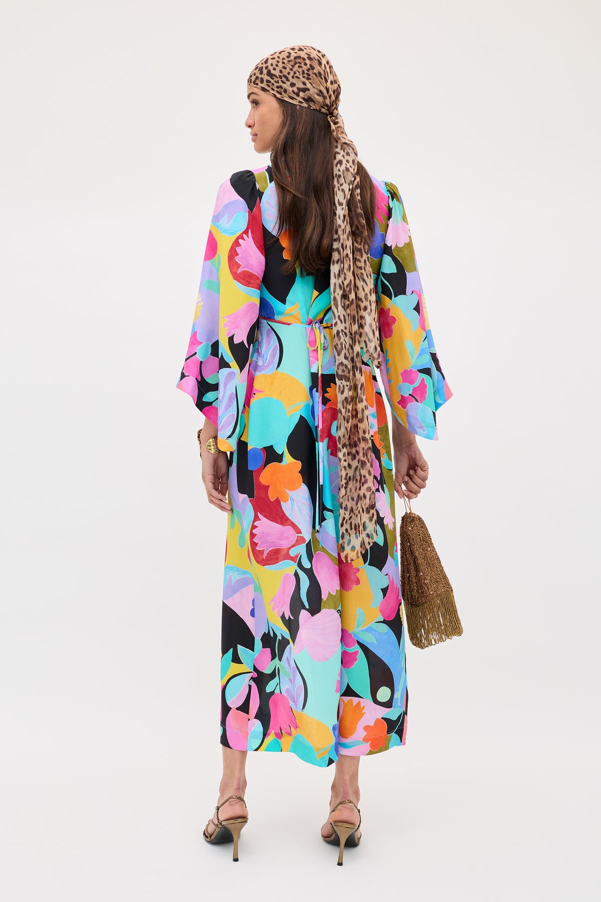 Facing away, a woman wears the Dahlia - Abstract Tulip Black silk crepe de chine dress and jacket set with dramatic sleeves, paired with strappy heels, a leopard-print headscarf, and a fringed gold handbag against a plain light background.