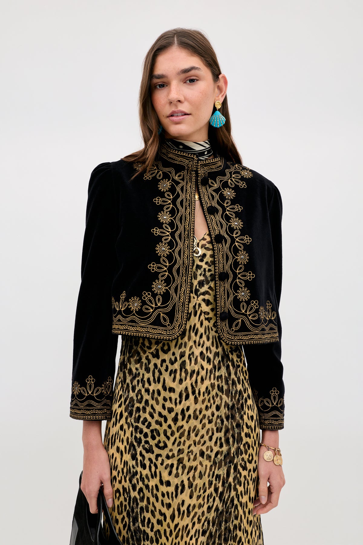 A woman wears the Denver - Soutache Black cotton-velvet jacket with gold embroidery over a leopard print dress, styled with blue earrings, a bracelet, and black clutch against a plain light background for a luxurious look.