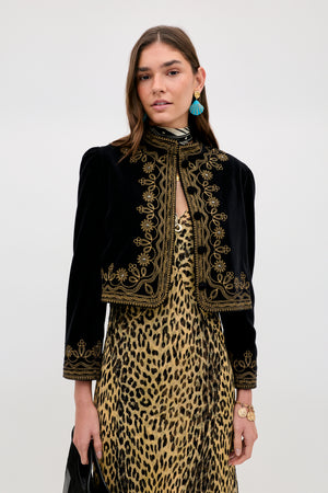 A woman wears the Denver - Soutache Black cotton-velvet jacket with gold embroidery over a leopard print dress, styled with blue earrings, a bracelet, and black clutch against a plain light background for a luxurious look.