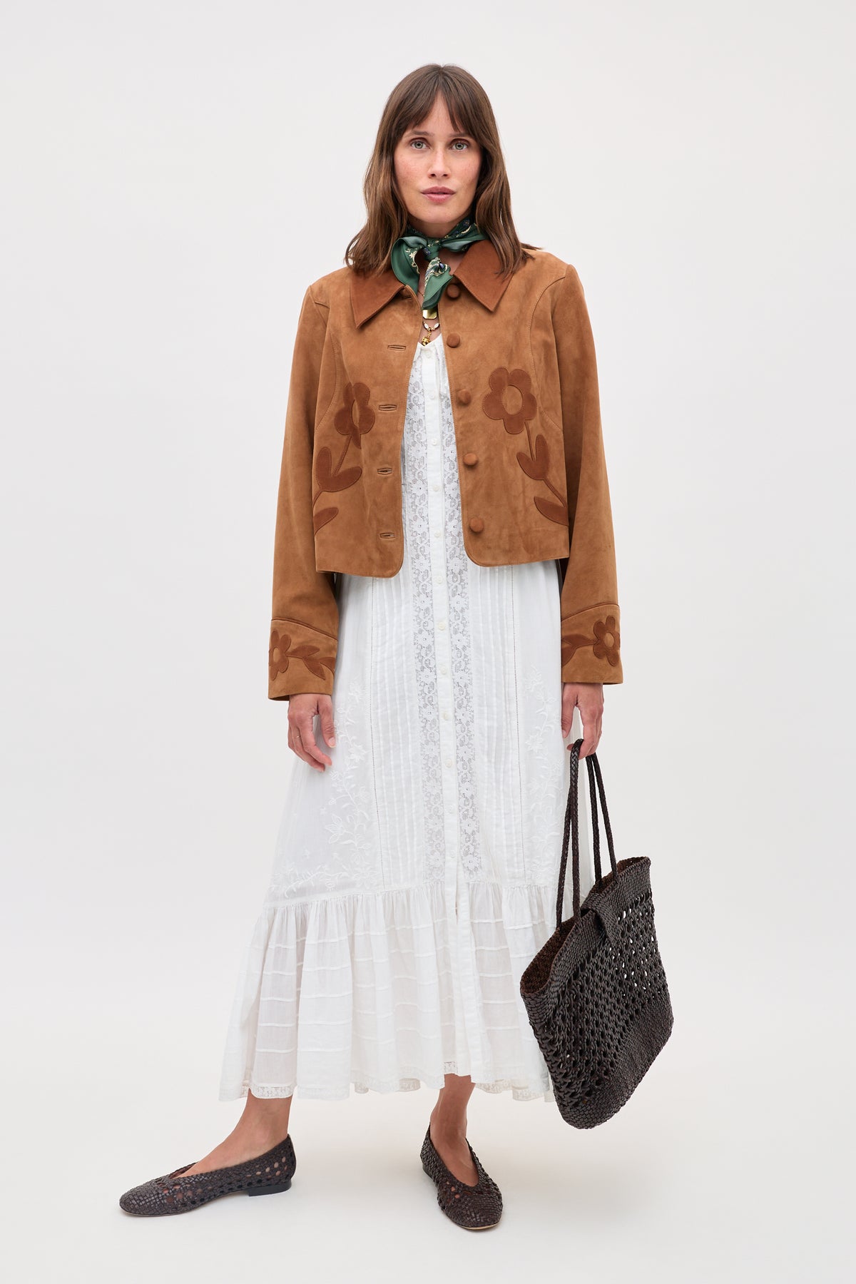 A woman stands against a plain background, wearing the Dionne - Retro Daisy brown suede jacket with daisy appliqué over a white lace dress, green scarf, brown woven shoes, and holding a black woven tote bag.