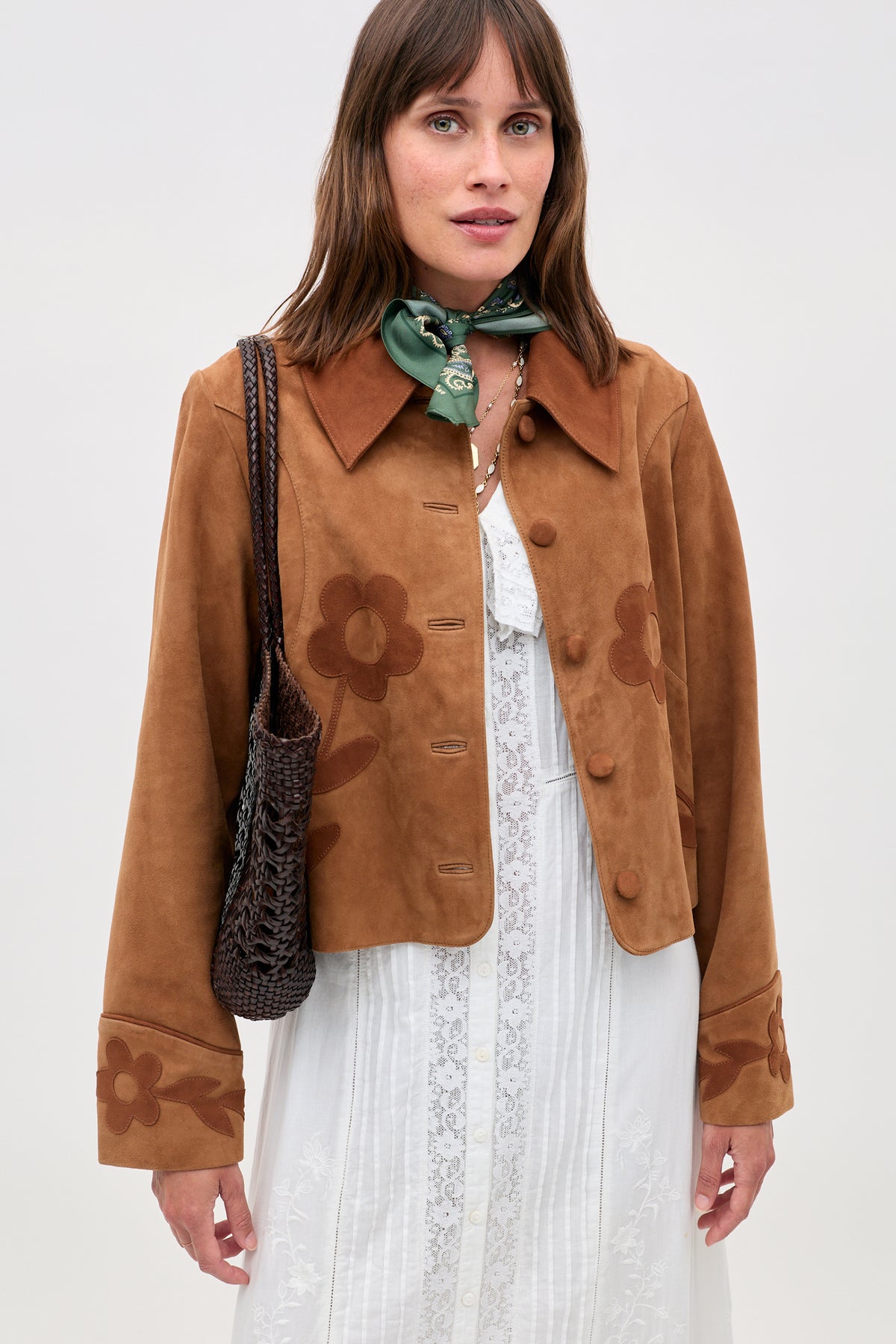 A woman in the Dionne - Retro Daisy brown suede jacket with daisy appliqué over a white lace dress, a green scarf, and holding a woven brown bag stands against a plain light background.