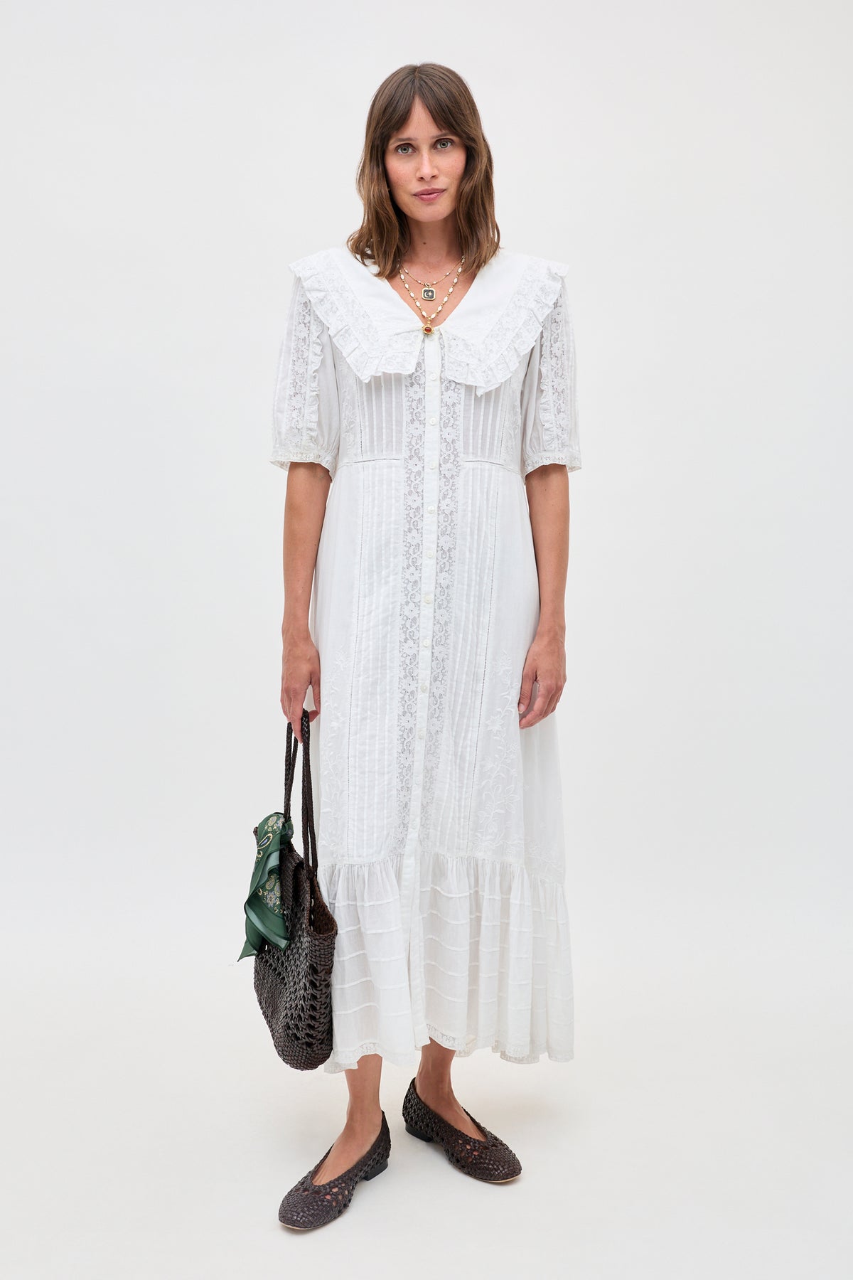 A woman wears the Edina - Pansy Embroidery White vintage-style, ankle-length dress with a large collar, carrying a woven brown bag and wearing dark woven flats against a plain background.
