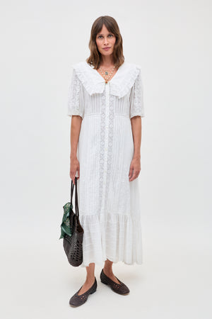 A woman wears the Edina - Pansy Embroidery White vintage-style, ankle-length dress with a large collar, carrying a woven brown bag and wearing dark woven flats against a plain background.