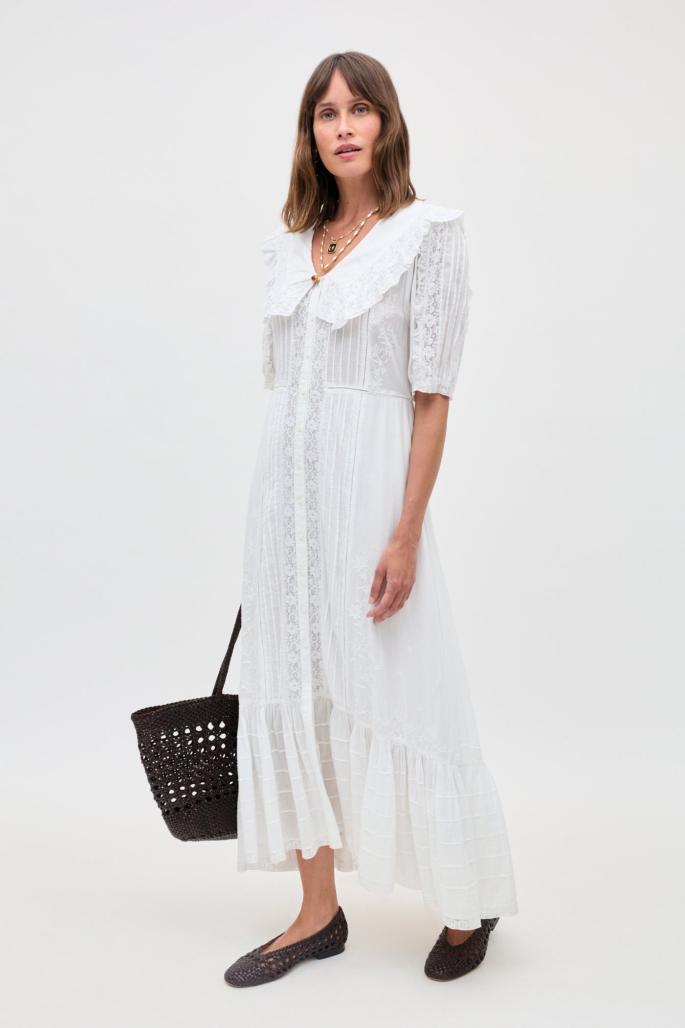 A woman wears the Edina - Pansy Embroidery White long lace blouse with a large collar, standing against a plain background, holding a black woven bag and wearing black woven flats.