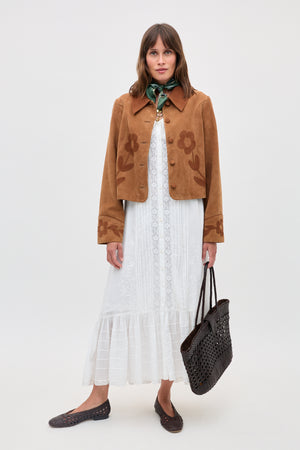 A woman models the Dionne - Retro Daisy tan suede jacket with daisy appliqué, styled over a white lace dress, green neck scarf, brown woven tote, and brown flats, standing against a plain light background.