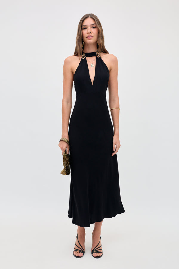 A woman stands against a plain background wearing the Eleanora - Black halter-neck dress with gold accents, paired with a small gold purse, black strappy heels, gold bracelets, and her long hair styled loose.