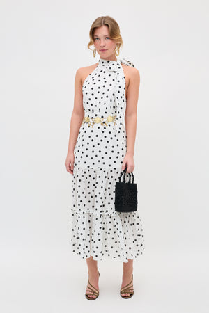 A woman in the Eleanor - Bohemia Spot Ivory dress—an ivory, polka-dot, sleeveless halter-neck midi—styled with a gold belt, strappy heels, and a black textured handbag stands against a plain white background.