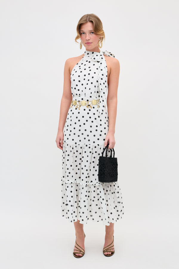 A woman in the Eleanor - Bohemia Spot Ivory dress—an ivory, polka-dot, sleeveless halter-neck midi—styled with a gold belt, strappy heels, and a black textured handbag stands against a plain white background.