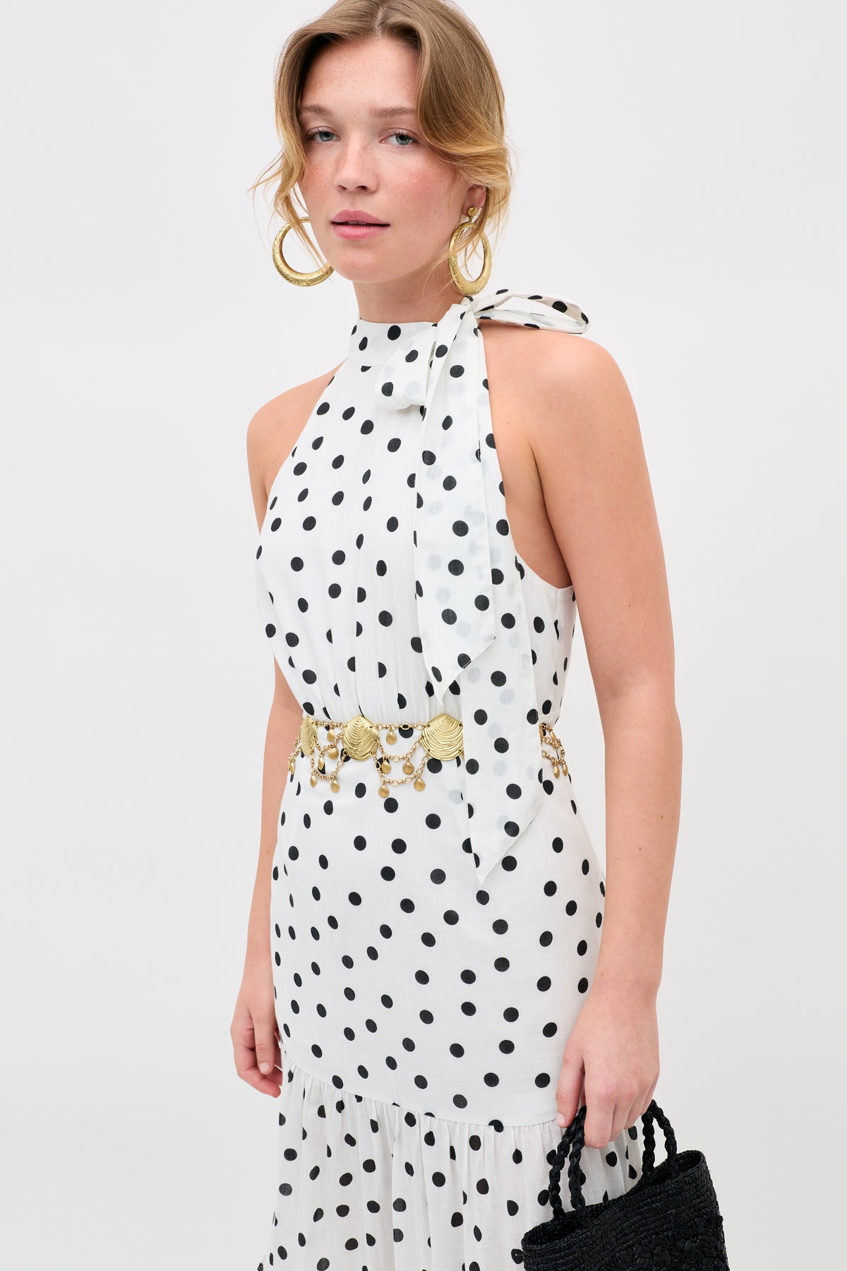 A woman wears Asia - Gold large gold hoop earrings, a sleeveless white halter dress with black polka dots, a gold belt, and holds a black woven handbag against a plain light background.