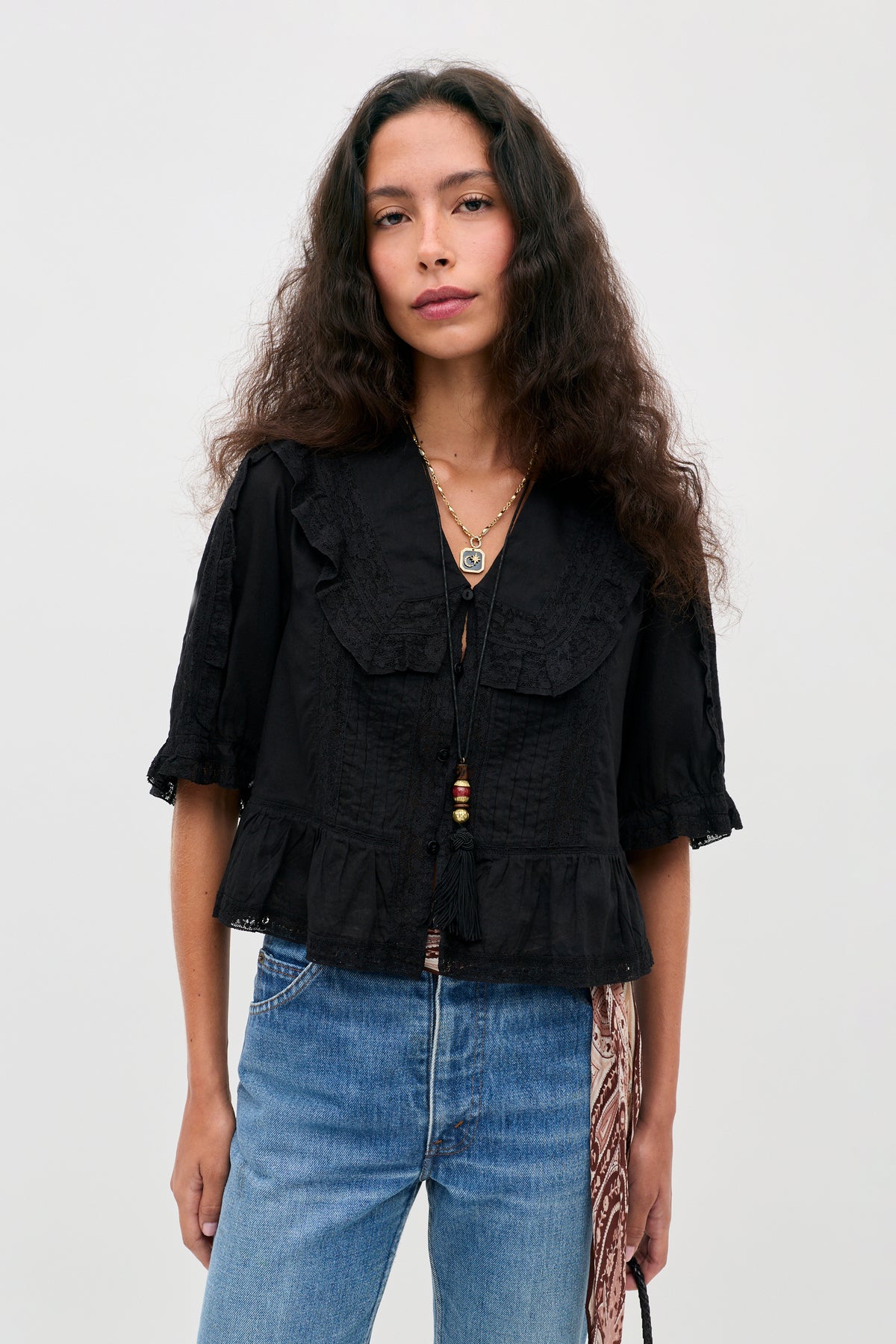 A woman with long wavy hair wears the “Ellery - Black,” a vintage-inspired black cotton button-down top with a delicate ruffled collar, paired with blue jeans and layered necklaces. She stands against a plain light background, holding a bag adorned with a patterned scarf.