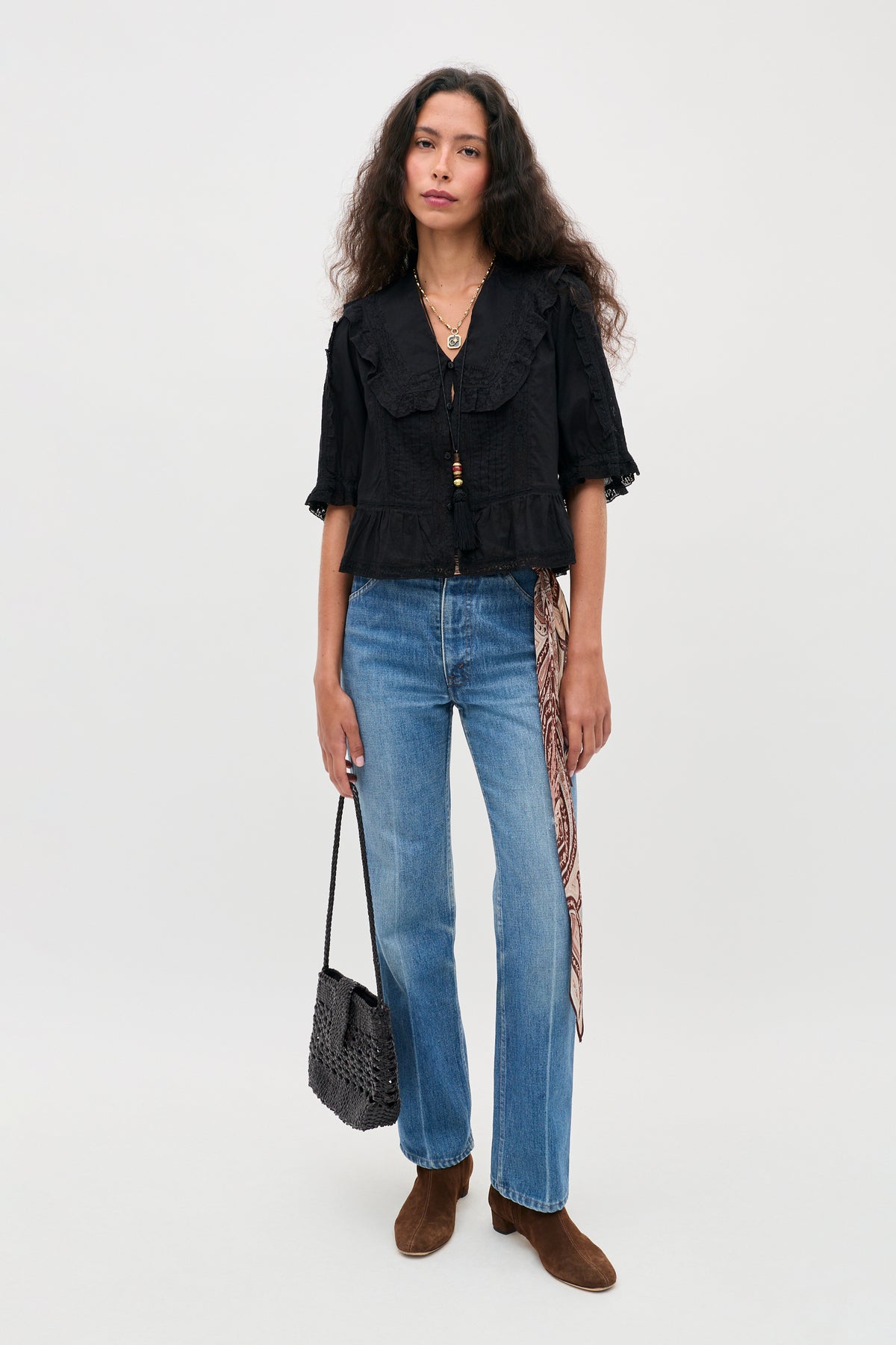A woman with long hair wears the Ellery - Black vintage inspired cotton button-down top featuring a ruffled collar, paired with blue jeans, brown boots, and a woven black bag. A patterned scarf is tied to her jeans as she stands against a plain light background.