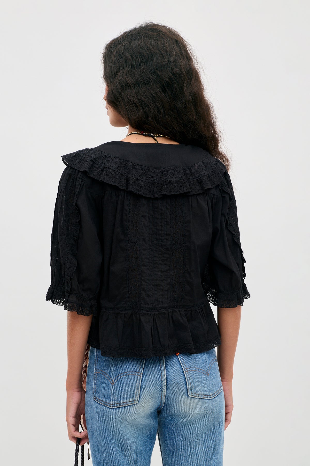 A person with long, wavy dark hair is shown from the back, wearing the Ellery - Black vintage-inspired cotton button-down top with a ruffled collar and three-quarter sleeves, paired with blue jeans. The background is plain light gray.