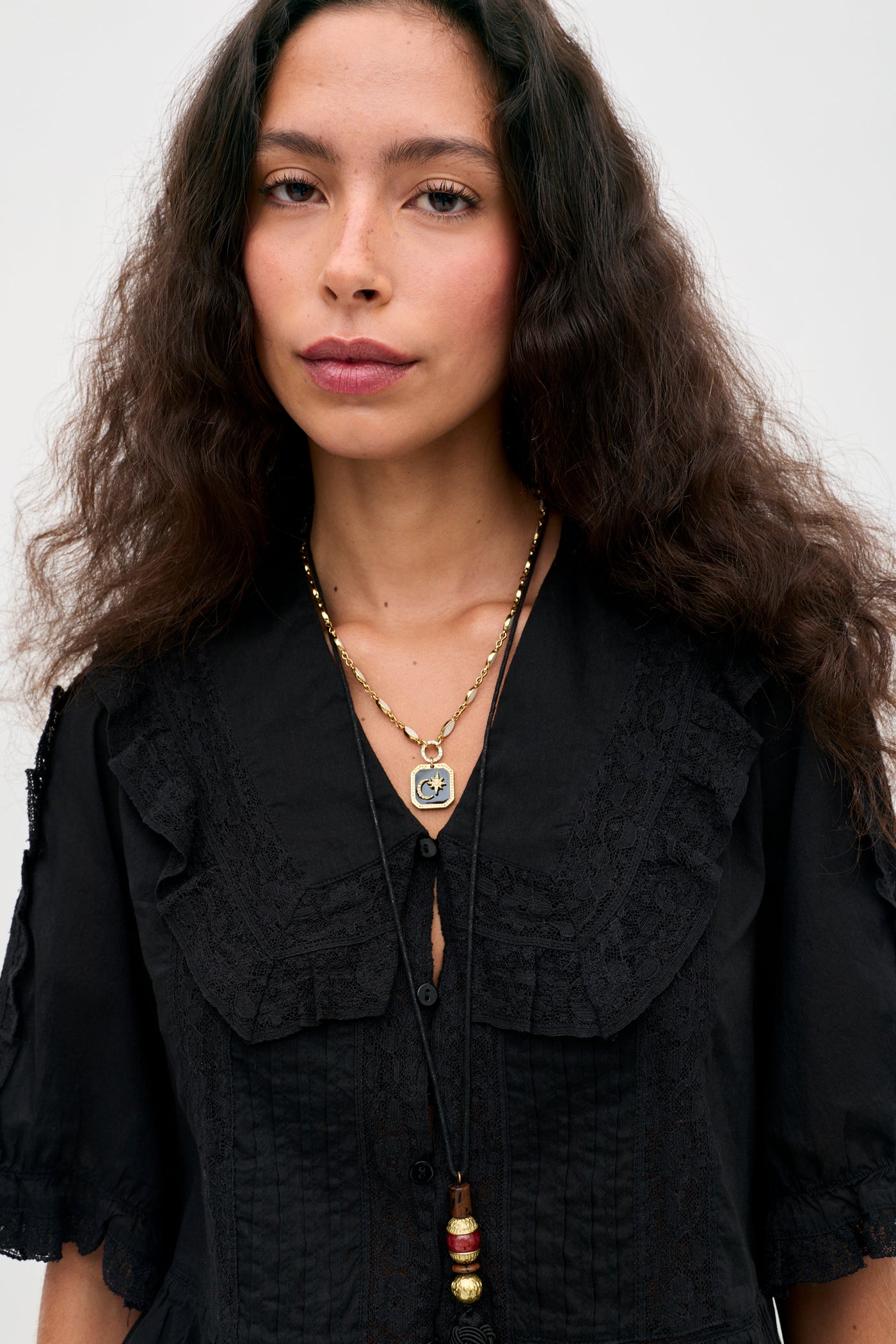 Wearing the “Ellery - Black” vintage-inspired cotton button-down top with ruffled details and a ruffled collar, a woman with long curly hair stands against a plain light background, pairing the look with layered pendant necklaces.