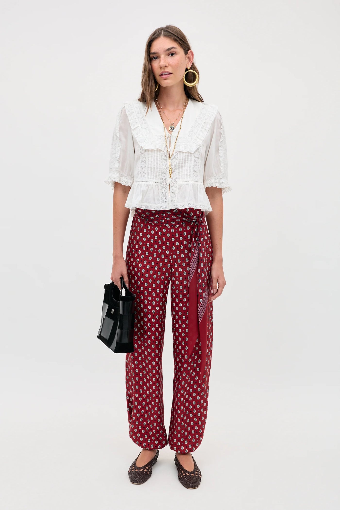A woman stands against a plain background wearing the Ellery - White blouse with a ruffled collar, styled with red patterned wide-leg pants, brown shoes, gold hoops, layered necklaces, and holding a black handbag.