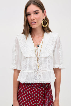 A woman wears the Ellery - White vintage-inspired sheer lace blouse with a ruffled collar and sleeves, paired with red patterned pants. She adds gold hoop earrings, layered necklaces, and carries a black bag.