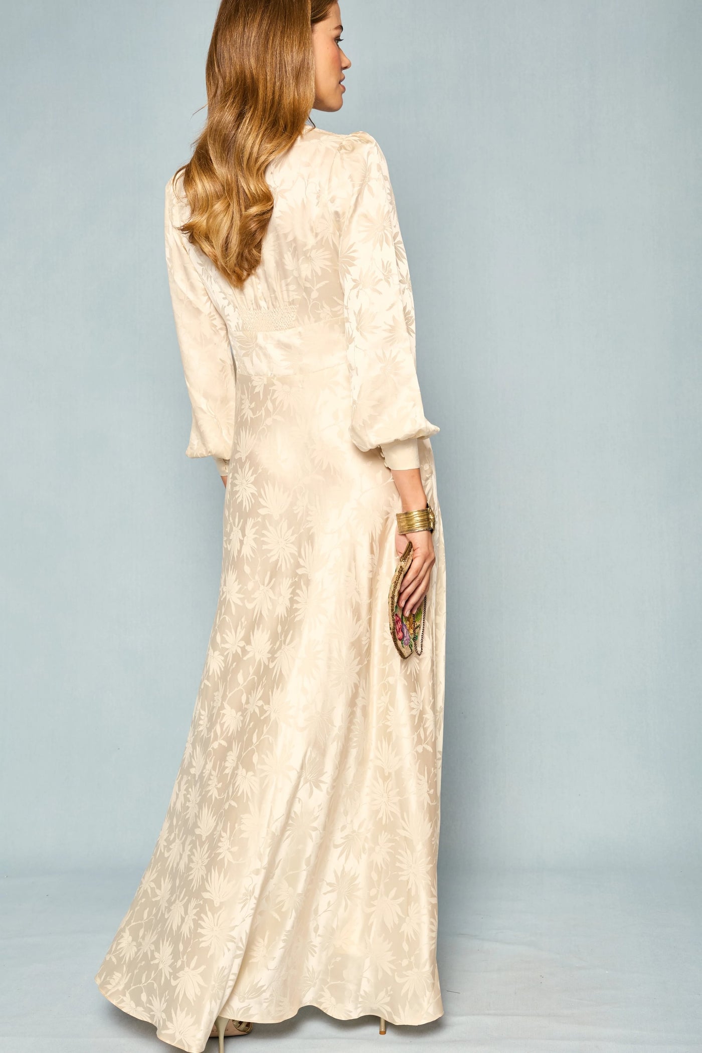 A woman with wavy hair wears the Emory - Daisy Jacquard Champagne, a long, cream-colored silk dress featuring delicate floral patterns. Holding a clutch, she stands facing away to reveal the elegant back details of the dress against a light blue background.
