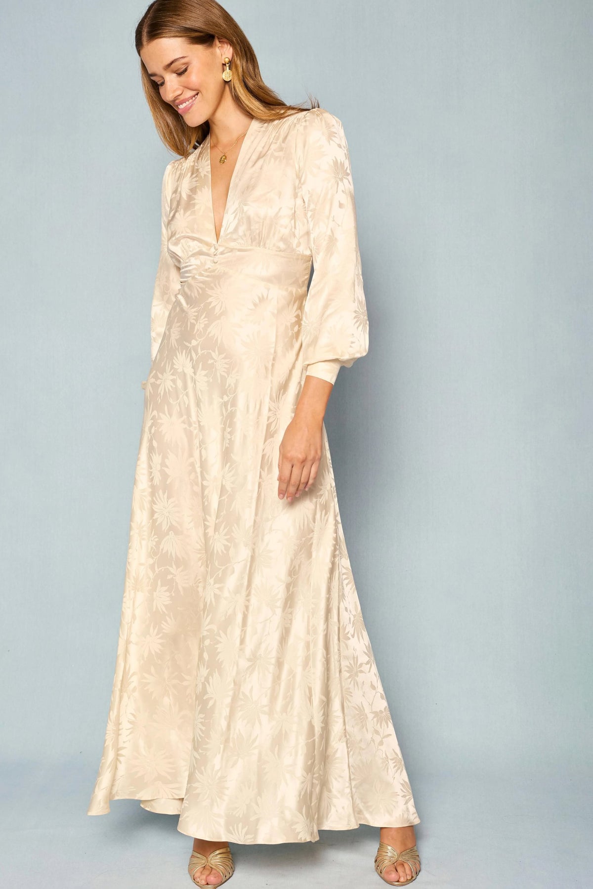 A woman stands against a plain light blue background, smiling and looking slightly downward, wearing the Emory - Daisy Jacquard Champagne dress—a long, cream-colored silk jacquard gown with floral patterns, long sleeves, and a deep V-neck.