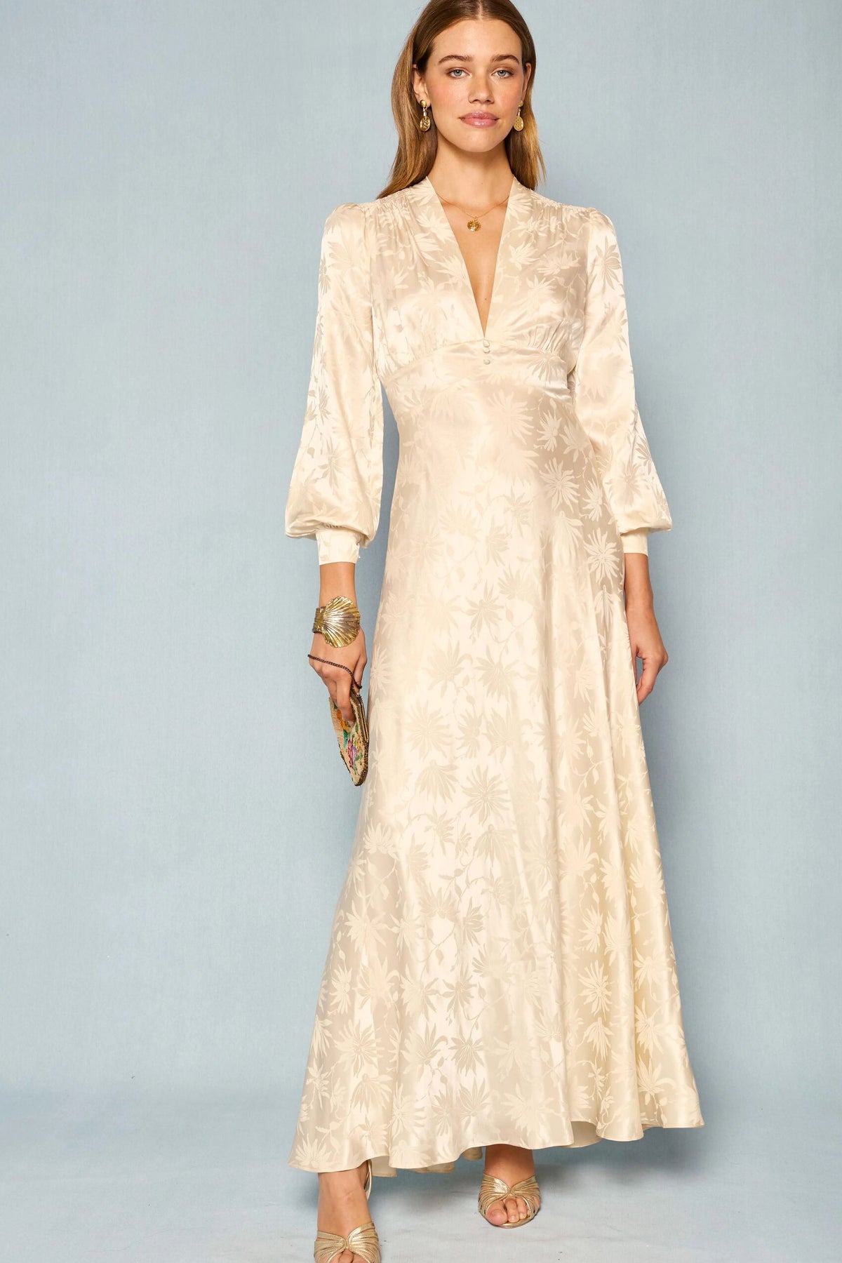 A woman wearing the Emory - Daisy Jacquard Champagne dress, a long cream-colored gown with a floral jacquard pattern and long sleeves reminiscent of a vintage wedding dress, stands against a light blue background, accessorized with a small clutch, gold sandals, and jewelry.