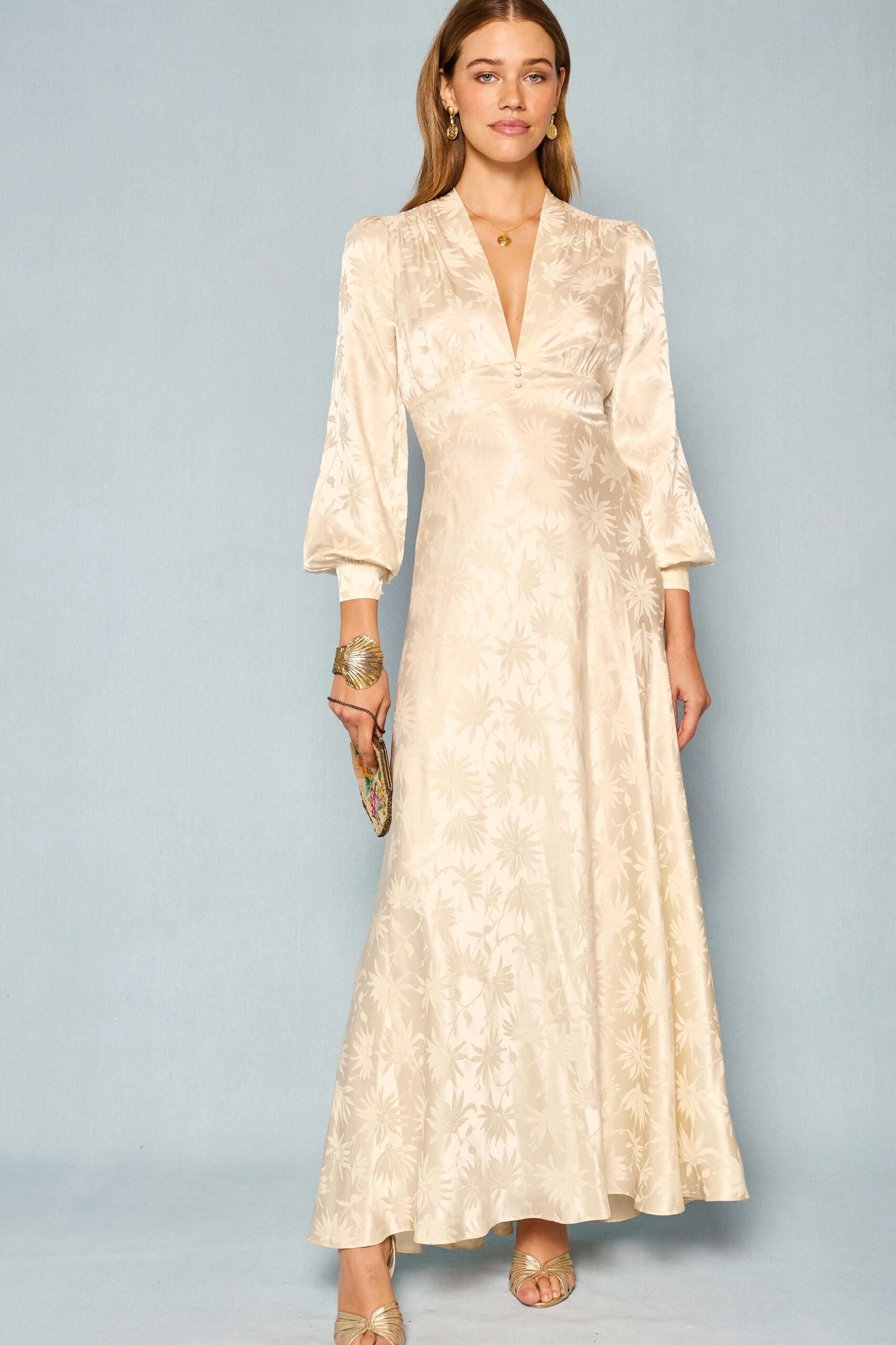 A woman wearing the Emory - Daisy Jacquard Champagne dress, a long cream-colored gown with a floral jacquard pattern and long sleeves reminiscent of a vintage wedding dress, stands against a light blue background, accessorized with a small clutch, gold sandals, and jewelry.