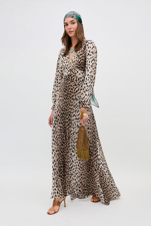 A woman wears the Emory - Antelope Spot Black dress, tan high-heeled sandals, a green patterned headscarf, and holds a gold fringed handbag as she stands against a plain white background.