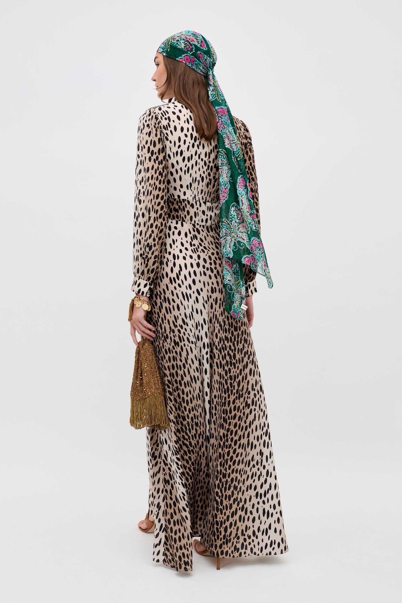 A woman stands facing away, wearing the Emory - Antelope Spot Black dress, brown heels, a green patterned headscarf, and holding a fringed gold handbag against a plain light background.