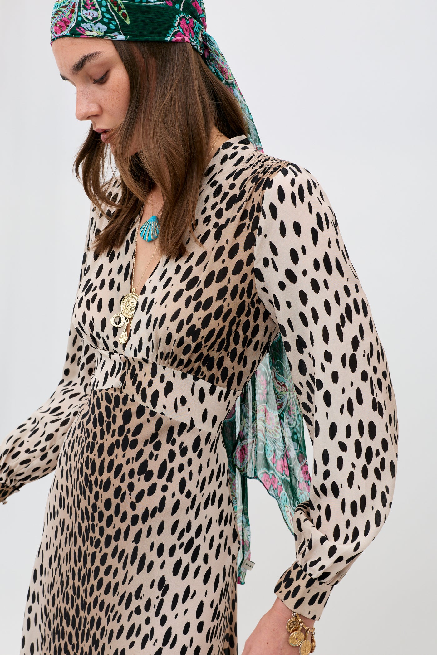 A woman in profile wears the Emory - Antelope Spot Black silk crepe de chine dress with gold buttons and long sleeves. She has brown hair, a floral headscarf, turquoise necklace, and gold bracelet against a plain white background.