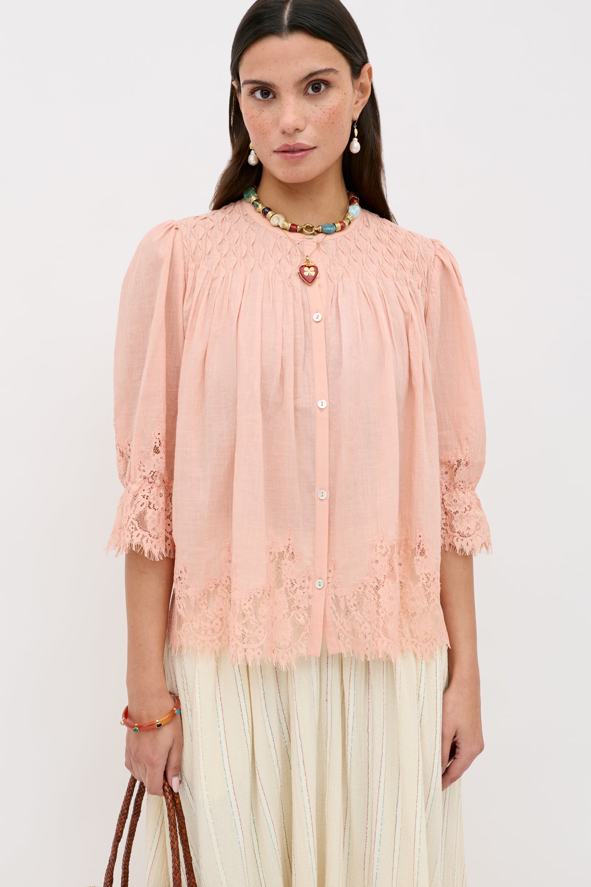 A woman wears the Erika Top in peach, a button-up blouse featuring lace trim and puffed sleeves, paired with a long cream skirt. She accessorizes with colorful jewelry and holds a brown bag, standing against a plain white background.