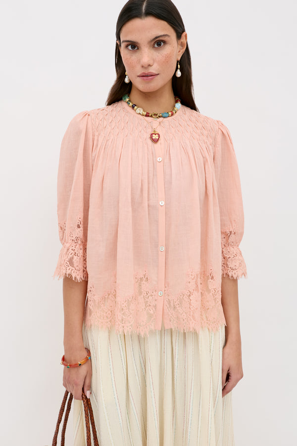 A woman wears the Erika Top in peach, a button-up blouse featuring lace trim and puffed sleeves, paired with a long cream skirt. She accessorizes with colorful jewelry and holds a brown bag, standing against a plain white background.