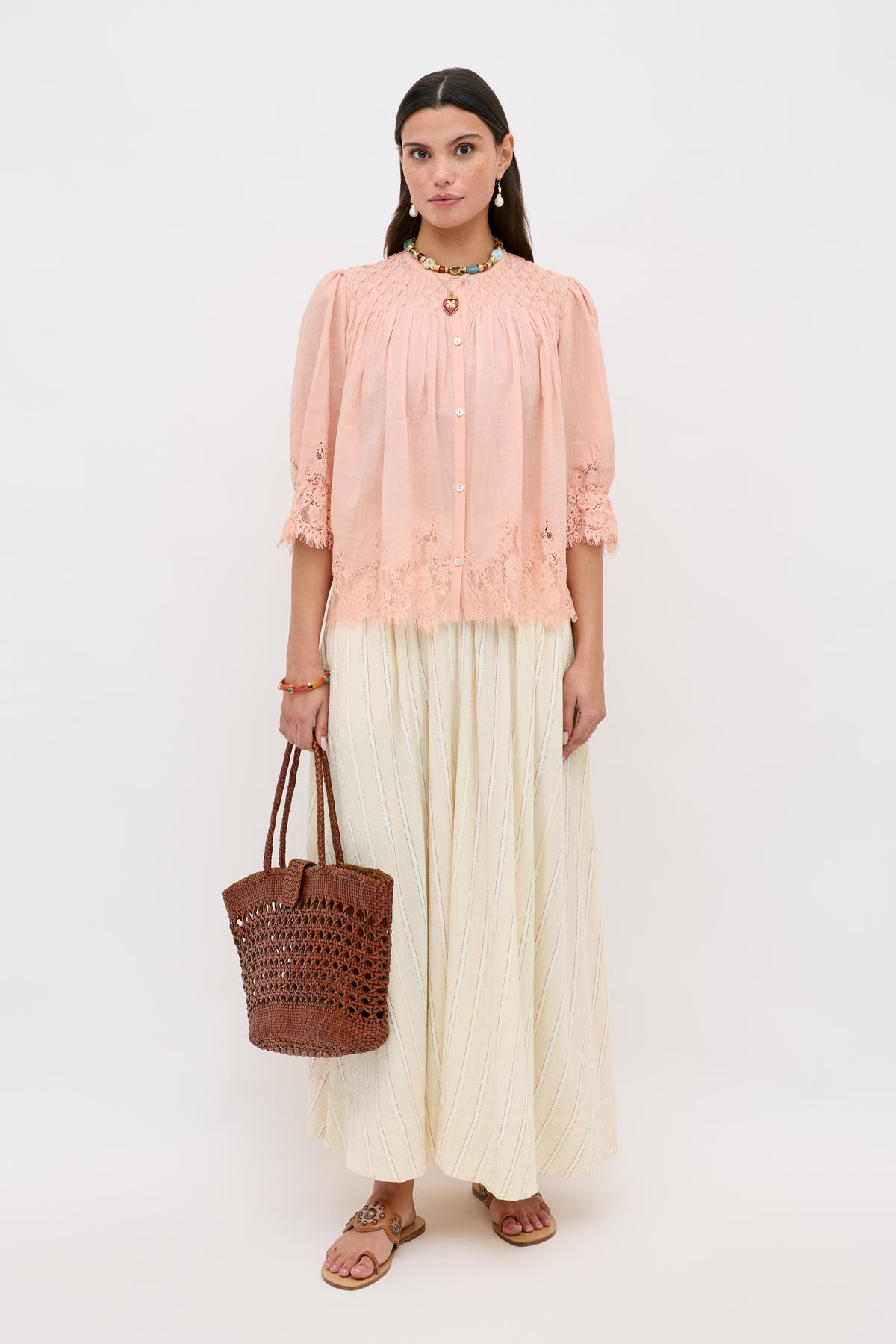 A woman stands against a plain background, wearing the erika top peach blouse, a cream maxi skirt, brown sandals, and holding a brown woven tote bag. She has long dark hair and wears simple jewelry.