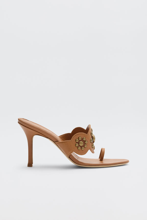 The Esmeray - Tan is an open-toe heeled sandal with a slim stiletto heel, wide cutout strap adorned with gold studs, and a thin toe strap, shown on a plain white background.