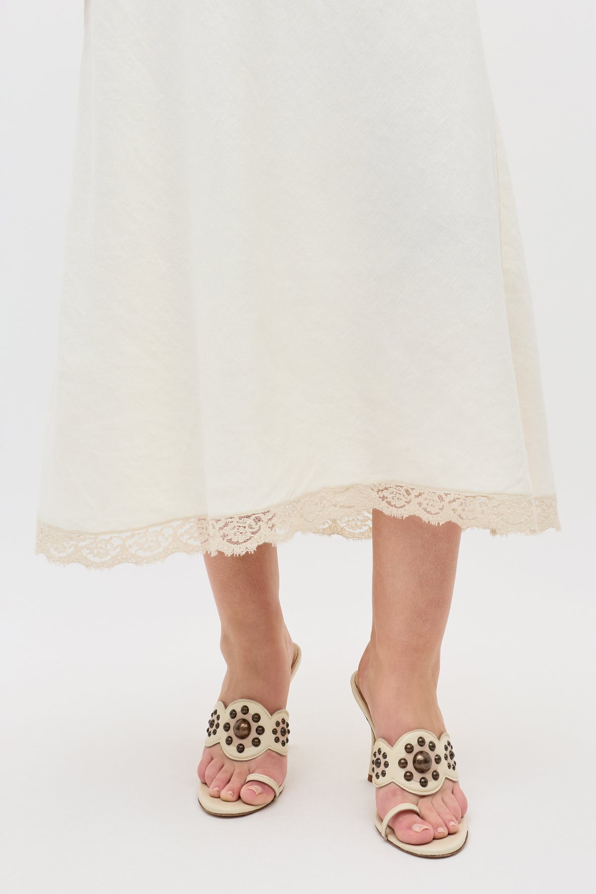 A person wearing the Esmeray - Cream long skirt with lace trim at the hem and beige heeled sandals with decorative studs, standing against a plain white background.