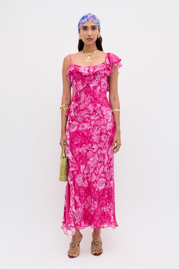 A person wearing the Ester - Cara Devore Magenta dress, paired with a blue and pink headscarf, gold jewelry, and holding a textured gold purse, stands against a plain white background.
