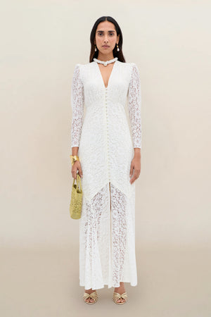 A woman stands against a neutral background wearing the Fleur - Ivory dress, a long white corded lace design with long sleeves and a subtle fit-and-flare silhouette. She completes the look with gold sandals, jewelry, and a gold handbag.