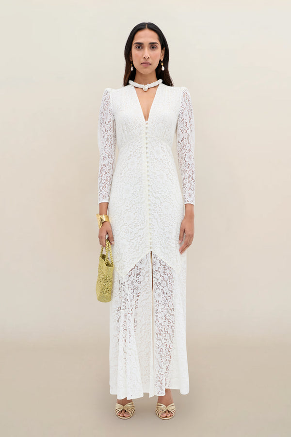 A woman stands against a neutral background wearing the Fleur - Ivory dress, a long white corded lace design with long sleeves and a subtle fit-and-flare silhouette. She completes the look with gold sandals, jewelry, and a gold handbag.